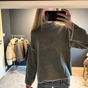 Grå stickad tröja från Zara - Mysig grå stickad tröja från Zara med hög hals och kontrastfärgade sömmar. Tröjan har en loose passform och långa ärmar, perfekt för lager-på-lager under kyliga dagar. Enkel och stilren design som funkar till det mesta.