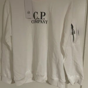 Vit sweatshirt från C.P. Company - Säljer en stilren vit sweatshirt från C.P. Company med svart logga på bröstet och ikonisk linsdetalj på ärmen. Tröjan har rund hals, långa ärmar och dragkedjeficka på ärmen. Perfekt för dig som gillar streetwear och snygga detaljer.