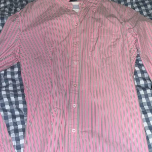 Rosa randig skjorta från Brooks Brothers, köpt på h&m - Säljer en klassisk rosa skjorta från Brooks Brothers men  köpt på h&m , med tunna gröna och vita ränder. Skjortan har lång ärm, bröstficka och knäppning framtill. Perfekt för dig som gillar färg och vill sticka ut lite extra.