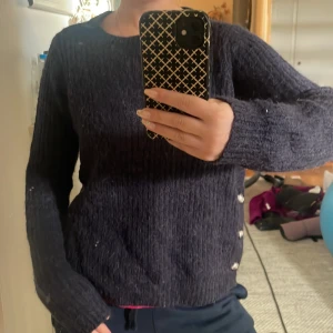 Mörkblå stickad tröja med knappar - Mysig mörkblå stickad tröja med rund halsringning och dekorativa knappar längs ena sidan. Perfekt för kyliga dagar och enkel att matcha med jeans eller kjol. Långärmad och i en klassisk, lite loose passform.