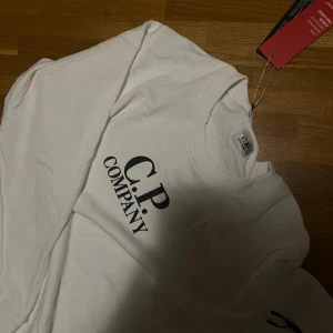 Vit sweatshirt från C.P. Company - Vit sweatshirt från C.P. Company med svart logga på bröstet och ikonisk linsdetalj på ärmen. Tröjan har rund hals, långa ärmar och en dragkedjeficka på ena ärmen. Perfekt för dig som gillar stilrena och trendiga plagg. Säljer denna då den inte riktigt är min stil, fick den av en släkting i present. Storlek M men skulle även säga att den funkar på personer som har storlek S vanligtvis, jag har S och den funkar på mig. Pris kan diskuteras vid snabbköp.