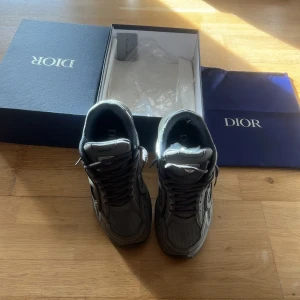 Grå Dior sneakers med box - Säljer ett par gråa Dior sneakers med snygga detaljer och klassisk logga på sidan. Skorna har snörning, rund tå och platt sula. Materialmix av skinn och textil ger en exklusiv känsla. Kommer med originalkartong och dustbag.