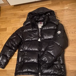 Svart Moncler pufferjacka med huva - Säljer en svart pufferjacka från Moncler i storlek M. Jackan har glansig yta, huva, dragkedja framtill och två fickor med dragkedja. Klassisk Moncler-logga på ärmen och quiltad design som håller värmen under kalla dagar.