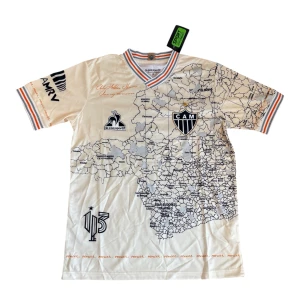 Atlético Mineiro fotbollströja Le Coq Sportif - Snygg vit Atlético Mineiro fotbollströja med kartmönster över hela framsidan, klubbmärke med stjärna på bröstet och broderade detaljer. Tröjan har korta ärmar och ribbade muddar med ränder i svart, orange och grått. Tillverkad i lätt och ventilerande polyester.