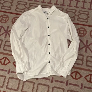 Vit skjorta från Stone Island L - Vit skjorta från Stone Island i storlek L. Klassisk design med svarta knappar framtill, lång ärm och krage. Skjortan är tillverkad i bomull och har en clean look som passar perfekt till jeans eller chinos.