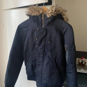 Mörkblå pufferjacka Polo Ralph Lauren - • Snygg mörkblå pufferjacka från Polo Ralph Lauren med fuskpäls på huvan, dragkedja och knappar framtill samt fickor på ärmen och sidan. Nyskick 