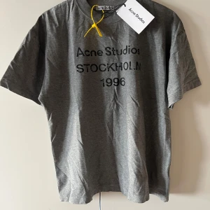 Grå Acne Studios t-shirt Stockholm - Snygg grå t-shirt från Acne Studios med svart tryck 'Stockholm 1996' på bröstet. Klassisk rund hals och korta ärmar. Tillverkad i mjuk bomull som känns skön mot huden. Perfekt för dig som gillar stilrena och trendiga plagg med streetkänsla.