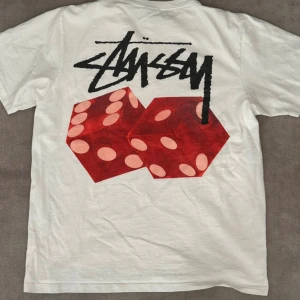 Vit Stüssy t-shirt med tärningar - Vit t-shirt från Stüssy i bomull med klassisk logga på bröstet och stor grafisk print med röda tärningar och svart Stüssy-tryck på ryggen. Modellen är kortärmad och har en rund halsringning. Perfekt för dig som gillar streetwear och snygga detaljer.