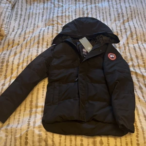 Svart dunjacka från Canada Goose M - Säljer en svart dunjacka från Canada Goose i storlek M. Jackan har huva, dragkedja och klassisk Canada Goose-logga på ärmen. Perfekt för kalla dagar med sin fluffiga och varma design. Snygg och stilren modell som håller dig varm hela vintern.