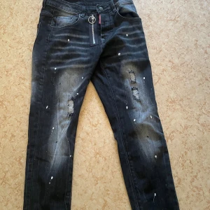 Svarta Dsquared2 jeans med dragkedja - Snygga svarta jeans från Dsquared2 med slitna detaljer, färgstänk och en cool dragkedja med ring framtill. Jeansen har en slim passform och är tillverkade i klassiskt denimtyg. Perfekta för dig som gillar streetstyle och vill sticka ut. Helt nya 