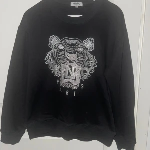 Svart Kenzo sweatshirt med tigerbrodyr - Svart sweatshirt från Kenzo med ikonisk silverbroderad tiger och logga på bröstet. Tröjan har rund halsringning, långa ärmar och en relaxed passform. Perfekt statement piece för dig som vill sticka ut med stil.