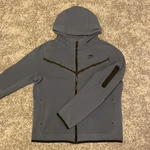 Mörk blå Nike tech - Nike tech i matt blått med hel dragkedja, huva och svarta detaljer. Techen har en sportig look med svarta linjer över bröstet och en praktisk ficka med dragkedja på ärmen. Perfekt för dig som gillar stilren och funktionell design.