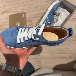 Blå mocka sneakers Christian Louboutin - Snygga blå sneakers från Christian Louboutin i mjuk mocka med vita snören och vit sula. Ikonisk röd undersida med Louboutin-logga. Perfekt för dig som vill sticka ut med exklusiva detaljer och stilren design.