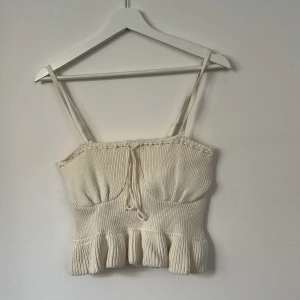 Stickad cremefärgad linnetopp Gina Tricot - Supersöt cremefärgad stickad linnetopp från Gina Tricot med smala axelband, volangkant nertill och knytning framtill. Toppen är ribbstickad och gjord i 100% bomull, perfekt för en chill och trendig look.