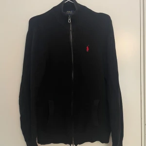 Ralph Lauren full zip - Elegant och tidlös tröja från Ralph Lauren, i en lyxig röd färg med matchande röd broderad logotyp på bröstet. Tillverkad i mjuk bomull av hög kvalitet som ger en bekväm och exklusiv känsla. Perfekt för både vardag och finare tillfällen.  Detaljer: 	•	Modell: Ralph Lauren Full Zip 	•	Färg: Röd 	•	Logotyp: Röd broderad pony 	•	Material: 100% bomull 	•	Passform: Normal / Regular fit 	•	Dragkedja framtill 	•	Ribbad mudd vid ärmar och midja 	•	Nypris: ca 2800 kr