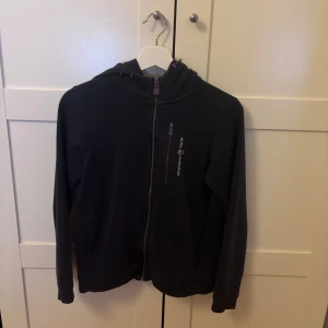 Svart hoodie från Sail Racing med dragkedja - Svart hoodie från Sail Racing med hel dragkedja framtill och en bröstficka med dragkedja. Tröjan har huva och långa ärmar, perfekt för dig som gillar en sportig och clean stil. Diskret logga på bröstet och på huvan.