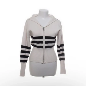 Randig hoodie med dragkedja från Zara - Vit och svart randig hoodie från Zara med dragkedja framtill. Tröjan är ribbstickad, har långa ärmar och huva. Tillverkad i en mix av viskos, polyester och polyamid. Snygg passform som markerar midjan och coola svarta ränder över bröst och ärmar.