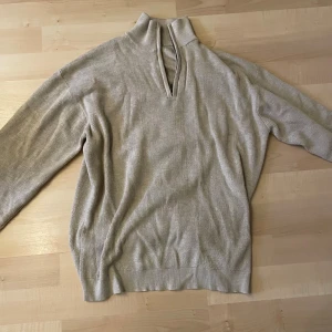 Beige ribbad stickad tröja med krage - Beige stickad tröja med ribbad struktur och hög krage med snygg v-ringad öppning framtill. Långärmad och i en loose passform, perfekt för lager-på-lager. Enkel och stilren design som passar till allt.