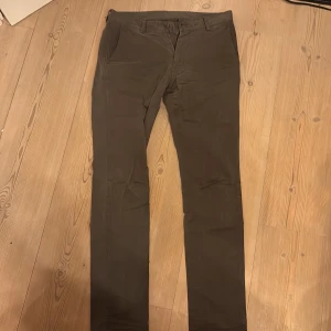 Bruna chinos från Paul & friends - Snygga bruna chinos från Paul & friends med klassisk rak passform. Byxorna har dragkedja, bälteshällor och bakfickor med knappar. Tillverkade i mjuk bomull med lite elastan för extra komfort. Perfekta för en clean och stilren look. Byxorna saknar knap vid gylfen men det ingår en knap som du kan sy på.