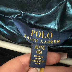Blå metallic pufferjacka Polo Ralph Lauren - Snygg blå metallic pufferjacka från Polo Ralph Lauren med huva och dragkedja. Jackan har broderad logga på bröstet och två fickor med dragkedja. Perfekt för kalla dagar när du vill sticka ut lite extra. Lite nött i vissa söner men varit där sen vi köpte den nästan tror det är färgen oxh sömnen som gör att det inte är lika mycket färg 