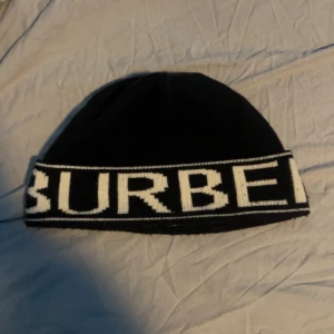 Svart mössa från Burberry - Svart stickad mössa från Burberry med vit logotyp runt hela kanten. Klassisk och stilren design med tydlig kontrast mellan svart och vitt. Perfekt accessoar för att lyfta din vinterstil.