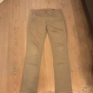 Slim chinos från Hansen & Jacob, beige - Beiga slim chinos från Hansen & Jacob med klassisk design, snedställda fickor fram och knappar bak. Tillverkade i bomull med lite stretch för skön passform. Perfekta för dig som gillar stilrena och bekväma byxor.