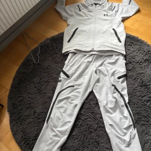 Grå träningsset från Under Armour - Säljer ett grått träningsset från Under Armour med jacka och byxor. Setet har svarta detaljer och dragkedjor på både jacka och byxor. Perfekt för dig som gillar sportig stil och vill ha något bekvämt och funktionellt.