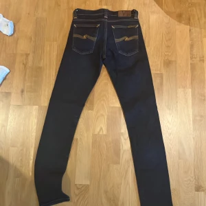 Mörkblå Nudie Jeans Co jeans W26 L34 - Snygga mörkblå jeans från Nudie Jeans Co med klassisk femficksdesign och kontrastsömmar. Modellen har raka ben och normal passform, med coola broderade detaljer på bakfickorna och läderpatch i midjan. Perfekt för dig som gillar stilrena jeans med edge. Storlek 26 W 32L. Modell long John 