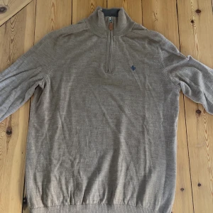 Beige stickad polotröja med half zip - Snygg beige stickad polotröja från Oscar Jacobson med half zip och broderad mörkblå logga på bröstet. Tröjan har lång ärm, ribbade muddar och hög krage. Perfekt för lager-på-lager under höst och vinter.
