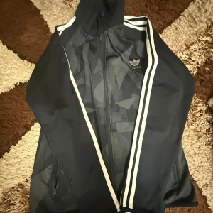 Svarta Adidas set - Svarta mjukisbyxor från Adidas med klassiska vita ränder längs sidorna och Adidas-logga på låret. Byxorna har elastisk midja och är tillverkade i ett mjukt, syntetiskt material som passar perfekt för träning eller chill. Snygg och sportig look. Helt nya 