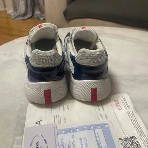 Prada sneakers i blått och vitt - Snygga Prada sneakers i marinblått och vitt med meshpaneler och blanka detaljer. Skorna har vita snören, rund tå och tydlig Prada-logga på plösen. Modellen av skorna heter Prada Cups