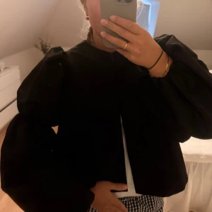  By Malina blus - Supersnygg svart jacka från By Malina i glansigt satinmaterial. Jackan har markerade puffärmar, rund halsringning och dekorativa knappar framtill. Perfekt statement-plagg för dig som vill sticka ut med en trendig och elegant look.