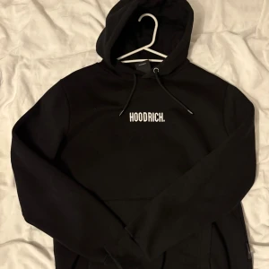 Svart hoodie från Hoodrich - Svart hoodie från Hoodrich med vit logga på bröstet. Klassisk huva med snörning och ribbade muddar. Mjuk och skön bomullsblandning, perfekt för chill eller streetwear. Snygga detaljer med Hoodrich-tryck på snörändarna. Använts 2-3 gånger pga för liten storlek, liten defekt vid snörningen vilket syns på bild 3. 