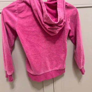 Rosa velourhoodie med dragkedja - Mysig rosa hoodie i mjuk velour med huva och dragkedja framtill. Tröjan har kängurufickor och ribbade muddar vid ärmslut och nederkant. Perfekt för chill dagar eller när du vill ha något bekvämt och snyggt.