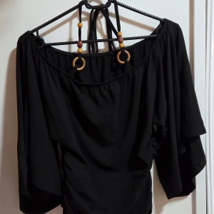 Svart offshoulder topp med trädetaljer - Svart offshoulder topp med vida ärmar och snygga träpärlor och ringar i axelbanden. Toppen har en lös passform och är tillverkad i mjukt bomullstyg. Perfekt för dig som gillar boho-vibbar och vill ha något unikt i garderoben.