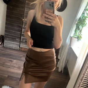 Brun skinnkjol med slits - Snygg brun kort skinnkjol med tight passform och en liten slits framtill. Kjolen har en stilren look och är gjord i ett glansigt material som ger en cool vibe. Perfekt för dig som vill sticka ut med en edgy och trendig stil.