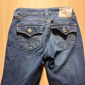 True Religion Becca Bootcut Jeans - Snygga blå jeans från True Religion, modell Becca Mid Rise Bootcut. Klassisk femficksdesign med markerade bakfickor och kontrastsömmar. Jeansen har medelhög midja och bootcut-ben, tillverkade i slitstarkt denim med coola detaljer på fickorna.