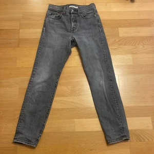 Levi's 501 grå jeans straight fit - Säljer ett par klassiska Levi's 501 jeans i snygg grå tvätt. Modellen har rak passform, fem fickor och knappgylf. Jeansen är tillverkade i slitstarkt denim och har den ikoniska läderpatchen bak i midjan. Har ett pytte litet hål på vänster knä men har sytt för. Så att det inte syns.146-152 men funkar för större