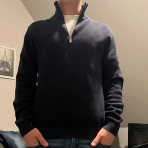 Mörkblå stickad tröja med half zip - Snygg mörkblå stickad tröja med half zip och hög krage. Tröjan har lång ärm och ribbade muddar vid ärmslut och nederkant. Perfekt för lager-på-lager och passar till jeans eller chinos.