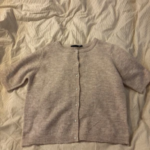 Beige kofta med pärlknappar från 157 - Superfin beige kofta från 157 med kort ärm och pärlknappar framtill. Mjuk stickad känsla och rund halsringning, perfekt att slänga över en topp för en chill look. Passar dig som gillar klassiska och enkla plagg med en söt twist.