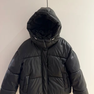 Svart pufferjacka från Bershka - Svart pufferjacka från Bershka i skinnimitation med dragkedja framtill och knappar. Jackan har en oversized vibe, stora fickor och quiltad look. Perfekt för kalla dagar när du vill ha en cool och trendig stil. Jackan är hel ny och har aldrig använt. 