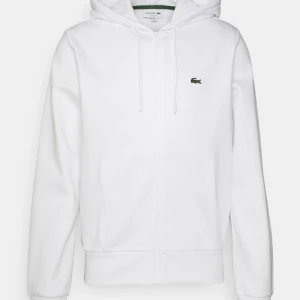 Vit hoodie från Lacoste med dragkedja - Säljer en clean vit hoodie från Lacoste med dragkedja och klassisk krokodil-logga på bröstet. Tröjan har huva med snörning, två stora fickor fram och är tillverkad i mjuk bomull. Perfekt för chill dagar eller när du vill hålla det stilrent. Köpt i Madrid för 180€ motsvarande 2000 kr endast använd 2/3 gånger 