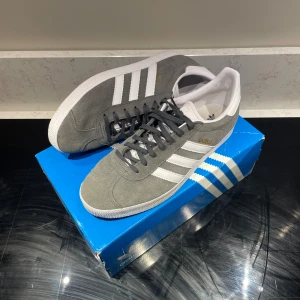 Adidas Gazelle grå/vita sneakers - Ett par nästan helt nya gazelle, de är använda 2 ggr, inköpta strax innan sommaren, säljer pga att de inte kommer till användning, kvitto finns!