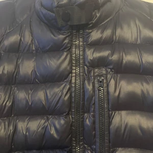 Svart pufferjacka från Saint Vincent - Snygg svart pufferjacka från Saint Vincent med glansig finish och markerad logga på ärmen. Jackan har hög krage, dragkedja framtill och en extra bröstficka med dragkedja. Perfekt för kalla dagar när du vill hålla stilen.