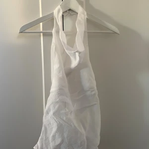 Vit omlott linnetopp med öppen rygg - Säljer en vit omlott linnetopp med halterneck och öppen rygg. Toppen har en snygg draperad front och knäpps i nacken. Materialet är lätt och luftigt. Det finns ett snöre om det önskas att knyta den så den sitter tajtare i midjan, jag använde inte de och gömde det (syntes inget alls)