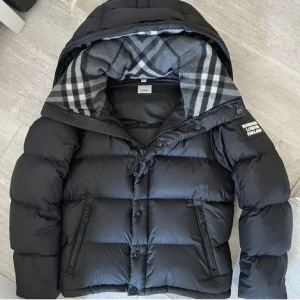 Svart pufferjacka från Burberry - Säljer en svart pufferjacka från Burberry med klassiskt rutigt foder i huvan. Jackan har dragkedja, knappar och två fickor framtill. Perfekt för kalla dagar och riktigt snygg streetstil. Märkespatch på ärmen och oversized känsla.