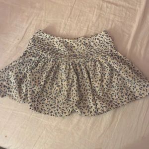 Blommig kortkjol från Gina Tricot - Supersöt kortkjol från Gina Tricot med små blå blommor på vit botten. Kjolen har smock i midjan för en bekväm passform och är gjord i ett lätt bomullstyg. Perfekt för en fräsch och somrig look!