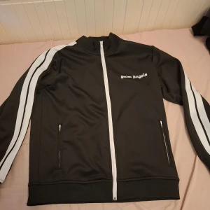 Palm Angels svart track jacket - Svart track jacket från Palm Angels med vita ränder längs ärmarna och vit dragkedja framtill. Jackan har ståkrage, två fickor med dragkedja och Palm Angels-logga på bröstet. Snygg och sportig stil, perfekt för streetwear.