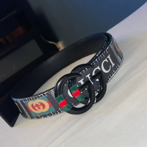 Svart Gucci bälte med GG-spänne - Säljer ett svart Gucci bälte med tydligt GG-spänne i svart, dekorerat med gröna och röda detaljer. Bältet har tryckt Gucci-logga och klassiska färgblock i grönt, rött och gult längs bandet. Tillverkat i skinn med snygga kontrastsömmar och bred design. Det finns fyra stycken spännhål. 