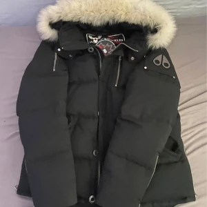 Svart Moose Knuckles dunjacka med päls - Säljer en svart dunjacka från Moose Knuckles med fluffig ljus päls runt huvan. Jackan har dragkedja, knappar och flera fickor med snygga detaljer. Perfekt för kalla dagar och har en riktigt schysst streetstil. Pris kan diskuteras :)